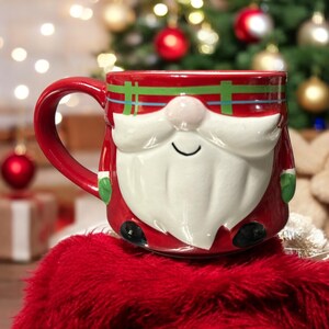 Peut inclure: Mug en c&eacute;ramique rouge en forme de P&egrave;re No&euml;l avec une barbe blanche et un chapeau &agrave; carreaux vert et rouge. La tasse est pos&eacute;e sur une surface rouge et duveteuse.
