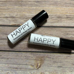 May include: Two small glass bottles with black rollerball tops. Both bottles have white labels with the word "HAPPY" and the text "Essential Oil Roller Bottle".