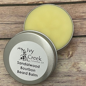 May include: A silver tin with a white label that reads "Ivy Creek, Columbus, GA, Sandalwood Bourbon Beard Balm". The tin is open and a yellow beard balm is visible inside.