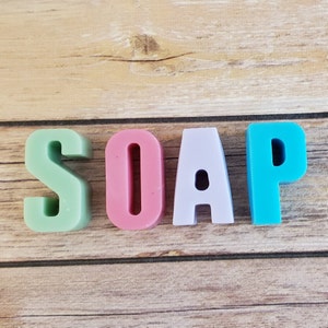 May include: The word "SOAP" spelled out in colorful, block-style letters. The letters are pastel shades of green, pink, lavender, and turquoise, arranged on a light brown wooden surface.