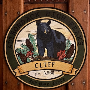 May include: A circular sign featuring a black bear illustration, pine branches, and red berries. The text reads ADIRONDACK HIGH PEAKS, CLIFF, elev. 3,960, and NEW YORK STATE. The design is set against a cream background with a dark green border.