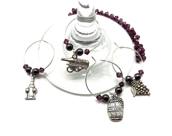 May include: Set of four wine glass charms with purple beads and silver charms. The charms include a wine bottle, a wine barrel, a bottle opener, and a bunch of grapes.
