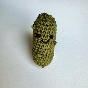 Op de afbeelding: Een groene gehaakte augurk met een glimlachend gezicht en roze wangen.