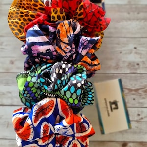 Könnte beinhalten: Ein Stapel bunter Seiden-Haargummis mit verschiedenen Mustern. Die Haargummis weisen lebendige Designs auf, darunter Leopardenmuster, Blumenmuster und abstrakte Formen in Rot, Orange, Blau und Grün. Im Hintergrund ist eine kleine Karte mit einem Logo zu sehen.