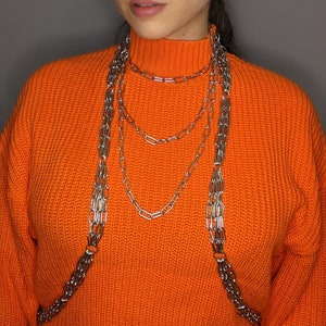 May include: A woman wearing a bright orange jumper with a silver chain necklace. The necklace is layered and has multiple strands.