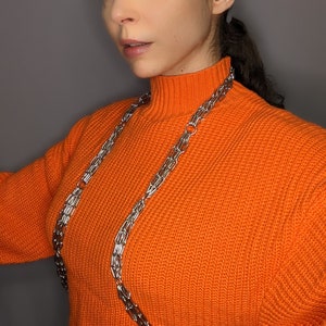 May include: A woman wearing a bright orange turtleneck sweater with a silver chain necklace. The chain is layered and extends down to her waist.