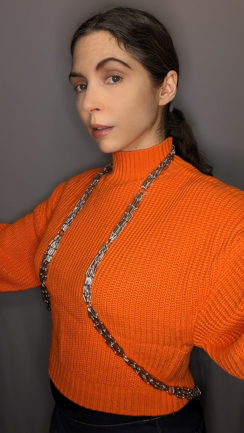 May include: A woman wearing a bright orange turtleneck sweater with a silver chain necklace. The chain is layered and extends down to her waist.