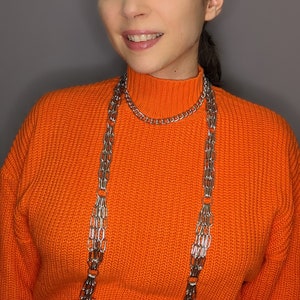 May include: A woman wearing an orange jumper with a silver chain body harness.