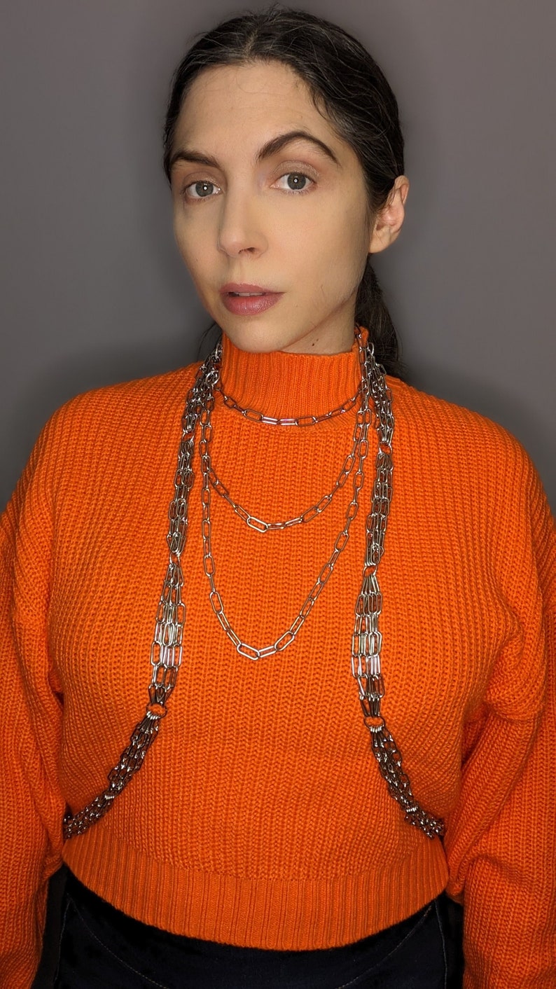 May include: A woman wearing a bright orange jumper with a silver chain necklace. The necklace is layered and has multiple strands.