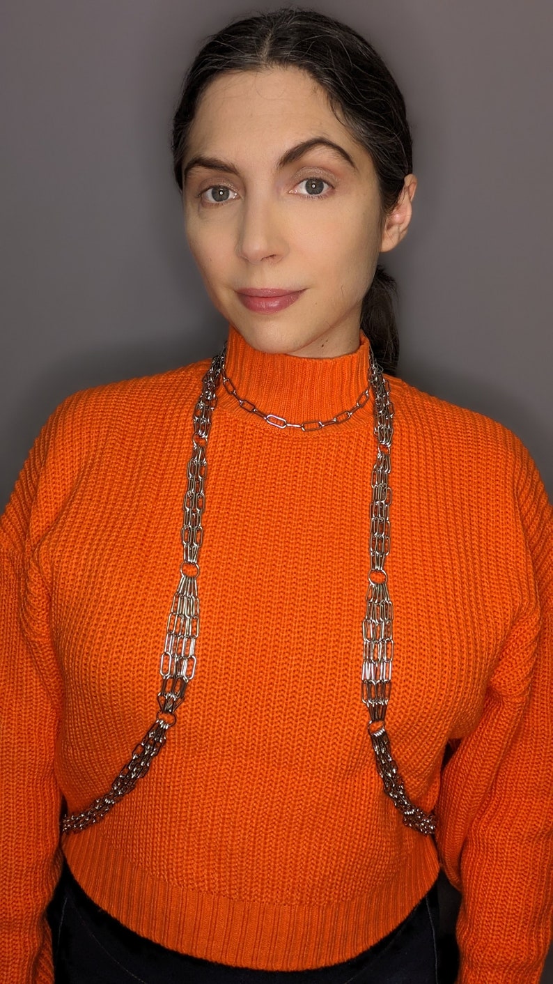 May include: A woman wearing an orange jumper with a silver chain necklace. The chain is layered and has a delicate design.