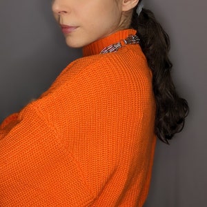 May include: A woman wearing a bright orange knitted jumper with a silver chain belt. The jumper has a high neck and a ribbed texture.