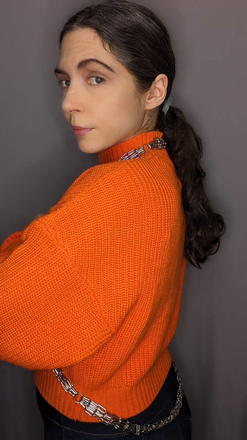 May include: A woman wearing a bright orange knitted jumper with a silver chain belt. The jumper has a high neck and a ribbed texture.