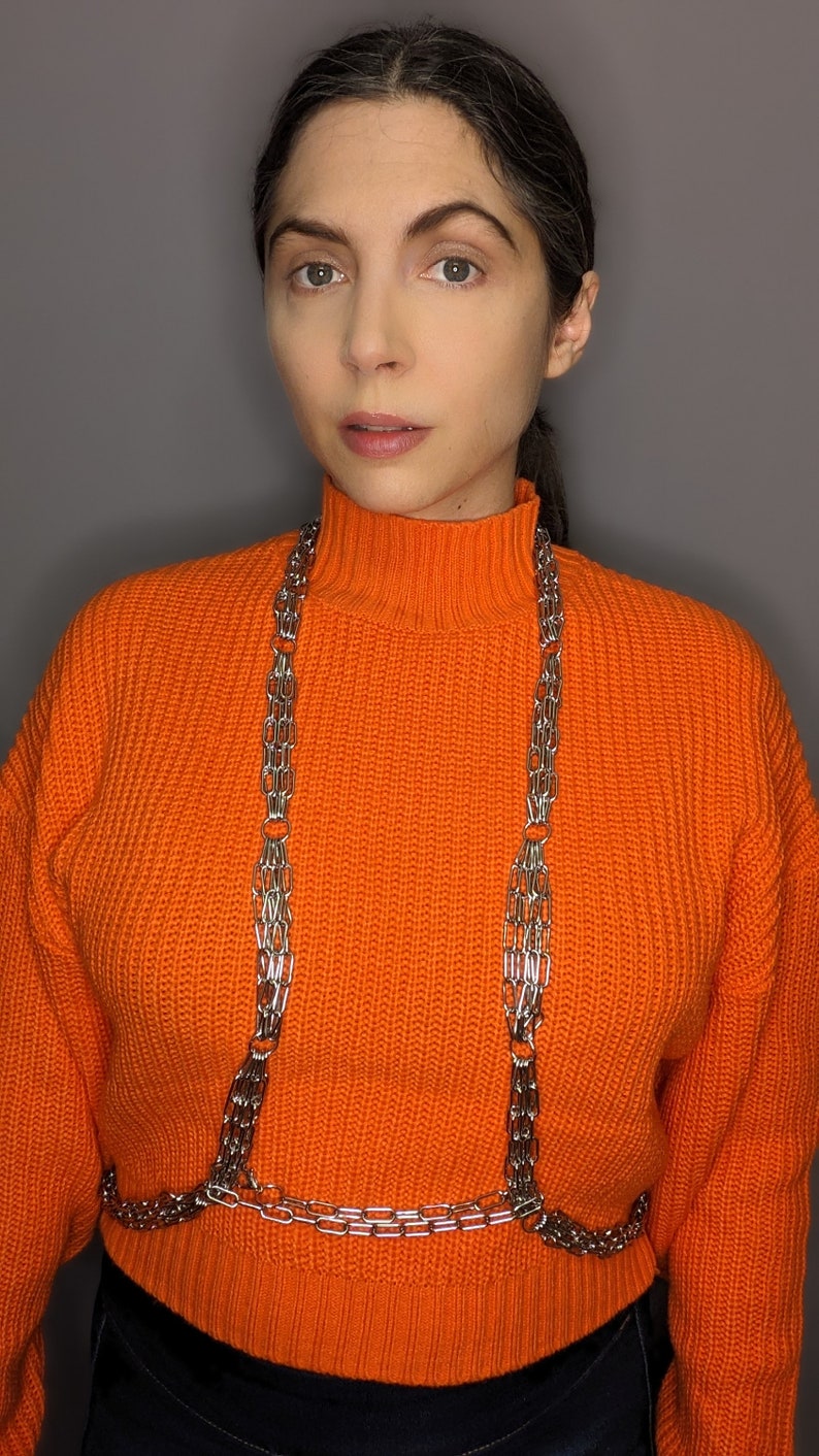 May include: A woman wearing a bright orange turtleneck sweater with a silver chain body harness.
