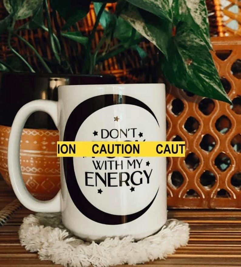 May include: White ceramic mug with a black crescent moon and stars design. The mug has a black handle and the text "Don't Caution With My Energy" printed on it.