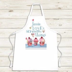 May include: White apron featuring a watercolour design of three cupcakes and the text "Jessie LOVES scrummy things." The cupcakes have pink, red, and yellow frosting. The apron has a blue gingham pattern and white lace trim.