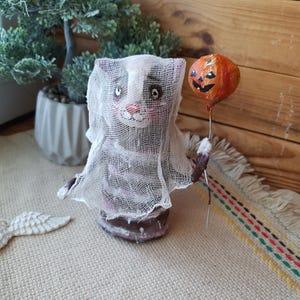 May include: A handmade Halloween ghost cat figurine with a white gauze-like fabric covering its body. The cat is holding an orange pumpkin balloon with a black jack-o'-lantern face.
