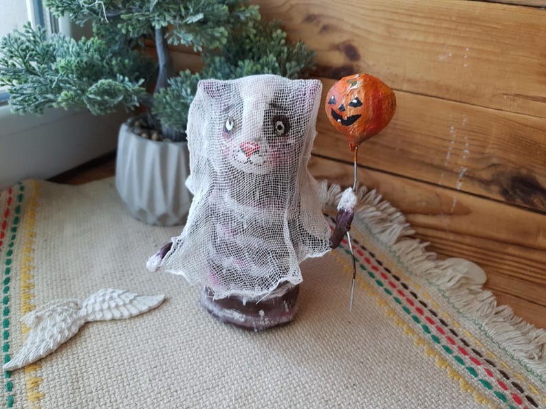 May include: A handmade Halloween ghost cat figurine with a white gauze-like fabric covering its body. The cat is holding an orange pumpkin balloon with a black jack-o'-lantern face.