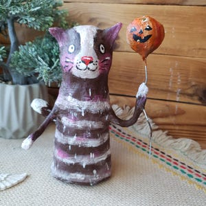 May include: A handmade paper mache cat figurine with white stripes, holding a small orange pumpkin balloon. The cat has a painted face with pink cheeks and a white nose.