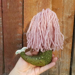 May include: A green mermaid doll with a pink yarn wig. The doll is holding its tail and has a green and gold scale pattern.