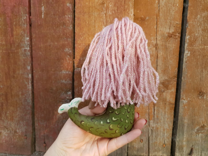 May include: A green mermaid doll with a pink yarn wig. The doll is holding its tail and has a green and gold scale pattern.