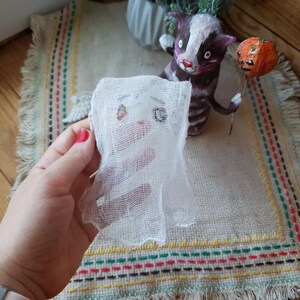 May include: A white gauze ghost with a drawn-on face, held in a hand. The ghost is in front of a handmade cat figurine holding a pumpkin.