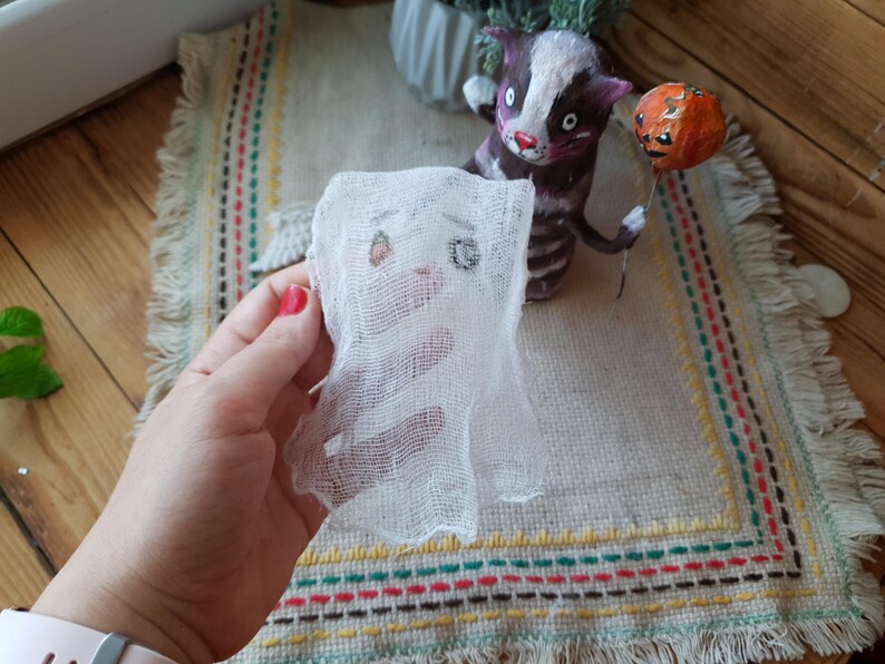 May include: A white gauze ghost with a drawn-on face, held in a hand. The ghost is in front of a handmade cat figurine holding a pumpkin.