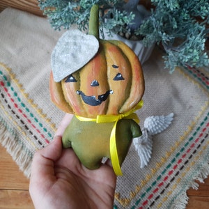 May include: A hand-painted, whimsical pumpkin figurine with a smiling face, a gray leaf-shaped hat, and a yellow ribbon. The pumpkin is primarily orange and green, with black accents for the eyes and mouth. A decorative item for fall or Halloween.