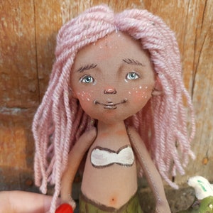 May include: A handmade doll with pink yarn hair, a white top, and a green tail. The doll has a painted face with brown eyes and a smile.
