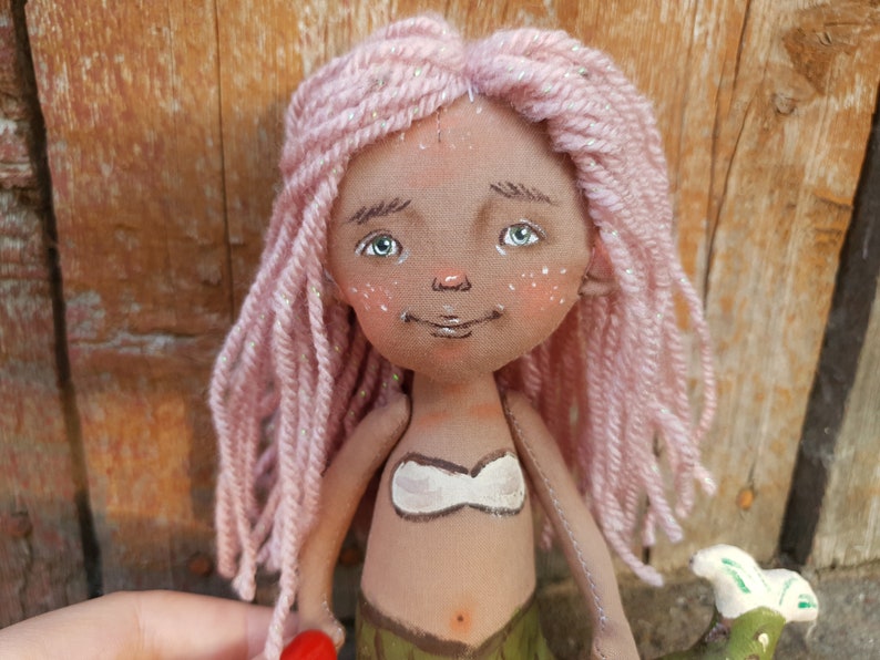 May include: A handmade doll with pink yarn hair, a white top, and a green tail. The doll has a painted face with brown eyes and a smile.