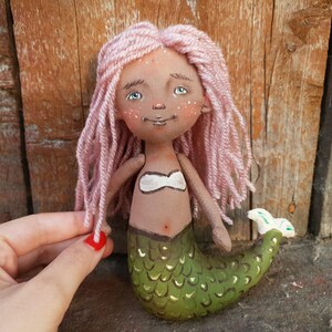 May include: A handmade doll with pink yarn hair, a brown face, and a green mermaid tail. The doll is wearing a white top and has a painted belly button.