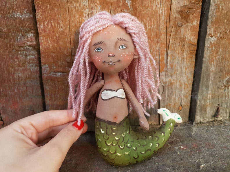 May include: A handmade doll with pink yarn hair, a brown face, and a green mermaid tail. The doll is wearing a white top and has a painted belly button.