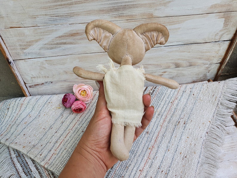 May include: A beige fabric doll with large ears and a simple white dress. The doll is being held by a hand.