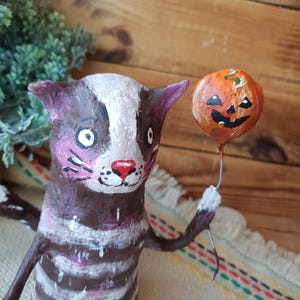 May include: A brown and white striped cat figurine with a pink nose and white eyes holds a small orange pumpkin balloon with a black jack-o'-lantern face.