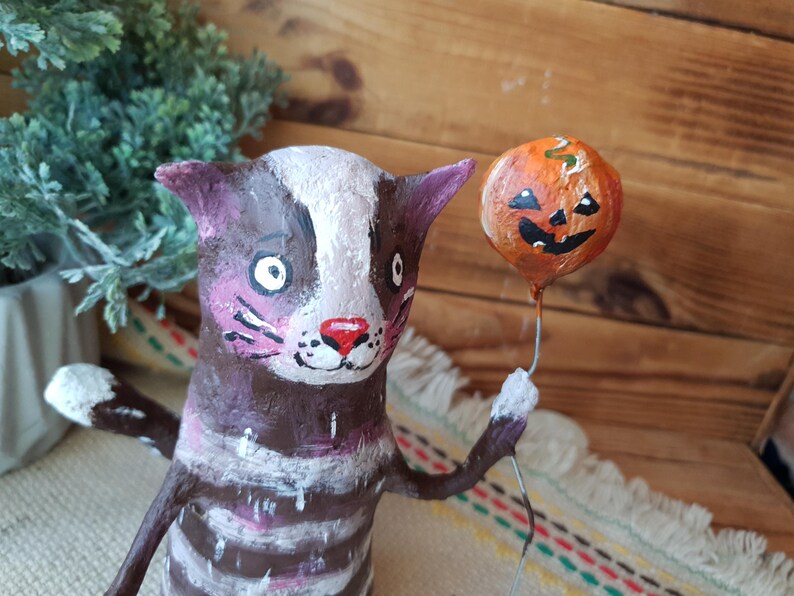 May include: A brown and white striped cat figurine with a pink nose and white eyes holds a small orange pumpkin balloon with a black jack-o'-lantern face.