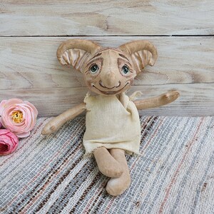 May include: A handmade plush toy elf with large ears, a beige body, and a white dress. The elf has painted eyes and a smiling mouth. The elf is sitting on a striped fabric.