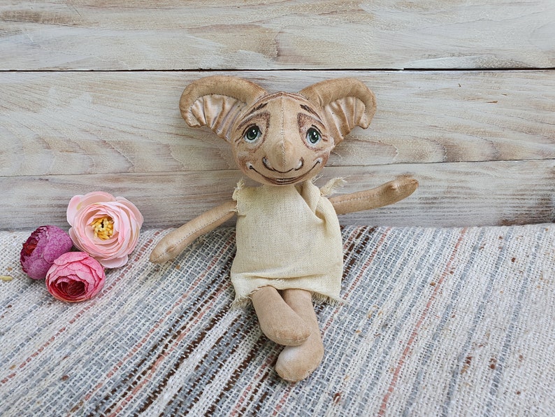 May include: A handmade plush toy elf with large ears, a beige body, and a white dress. The elf has painted eyes and a smiling mouth. The elf is sitting on a striped fabric.