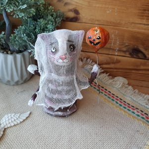 May include: A handmade Halloween cat figurine wearing a white gauze sheet and holding a small orange pumpkin balloon. The cat has black and white stripes and a painted face.