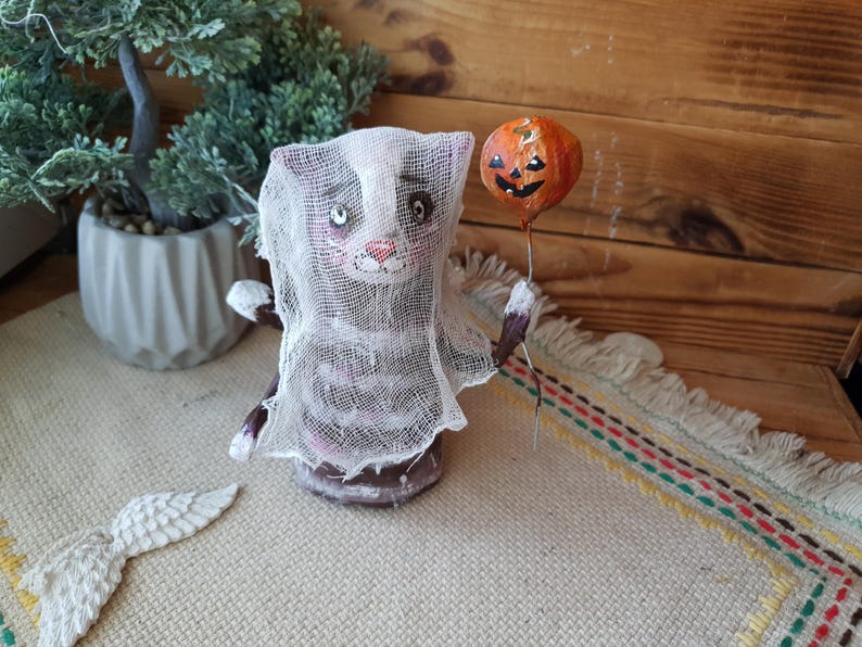May include: A handmade Halloween cat figurine wearing a white gauze sheet and holding a small orange pumpkin balloon. The cat has black and white stripes and a painted face.