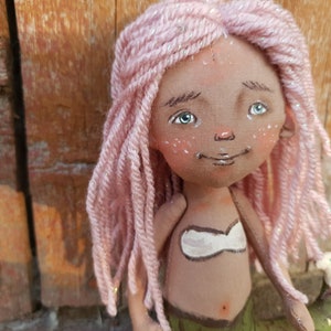 May include: A handmade doll with pink yarn hair, brown skin, and a painted green mermaid tail. The doll is wearing a white top and has a painted smile.