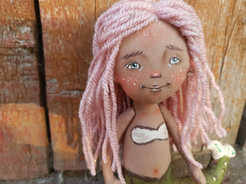 May include: A handmade doll with pink yarn hair, brown skin, and a painted green mermaid tail. The doll is wearing a white top and has a painted smile.
