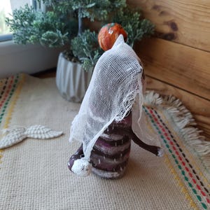 May include: A handmade doll with a brown and white striped dress, a white veil, and a small orange pumpkin on its head. The doll is standing on a beige rug with colorful stripes.