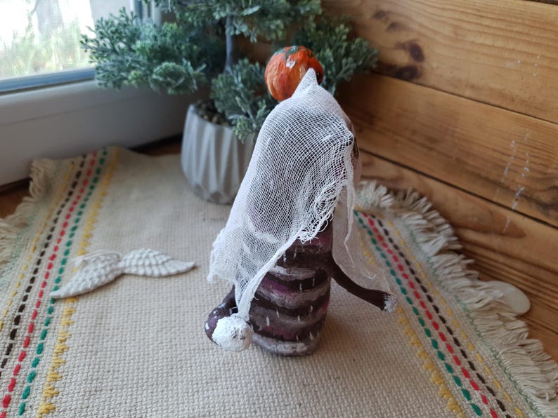 May include: A handmade doll with a brown and white striped dress, a white veil, and a small orange pumpkin on its head. The doll is standing on a beige rug with colorful stripes.