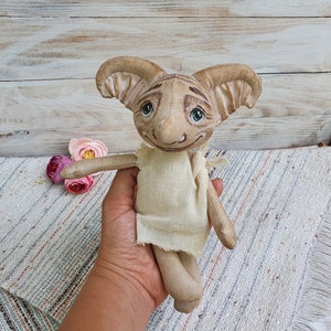May include: A handmade plush toy with large ears, a beige body, and a painted face. The toy has a light brown and beige striped pattern on its face and large, green eyes.