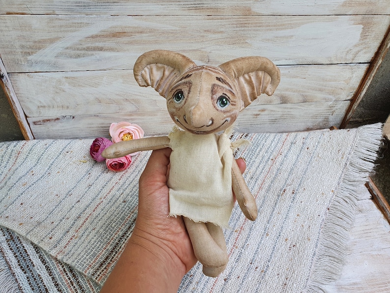 May include: A handmade plush toy with large ears, a beige body, and a painted face. The toy has a light brown and beige striped pattern on its face and large, green eyes.