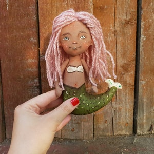 May include: Handmade doll with pink yarn hair, brown skin, and a green mermaid tail. The doll is wearing a white seashell bra.