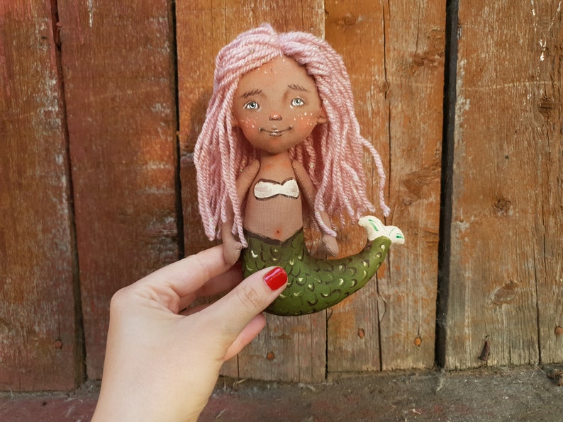 May include: Handmade doll with pink yarn hair, brown skin, and a green mermaid tail. The doll is wearing a white seashell bra.