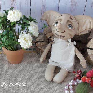 Peut inclure: Une poupée artisanale représentant un lutin de maison avec de grandes oreilles, une robe blanche et un visage peint. La poupée est assise sur une surface en bois avec une plante en pot et un collier de perles au premier plan.
