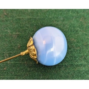 May include: A blue glass pin with a gold metal base. The pin is round and has a swirl pattern.