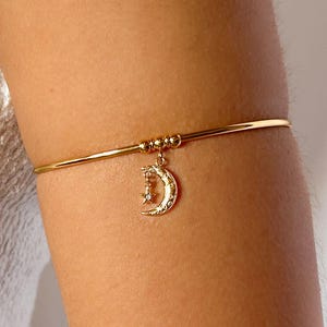 May include: Gold-toned arm cuff featuring a crescent moon charm with a star accent. The arm cuff is a thin, delicate band with small beaded details. The moon charm hangs from the band, adding a celestial touch to the jewelry.