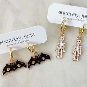 May include: Two pairs of gold hoop earrings with charms. One pair features a black bat with white eyes and a cross, and the other pair features a white skeleton.